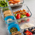 Side view of 3 plant based bistro boxes loaded with chickpeas, edamame, rice balls, carrots, cucumbers, tomatoes, berries and plant based yogurt.