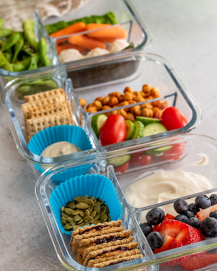 Side view of 3 plant based bistro boxes loaded with chickpeas, edamame, rice balls, carrots, cucumbers, tomatoes, berries and plant based yogurt.