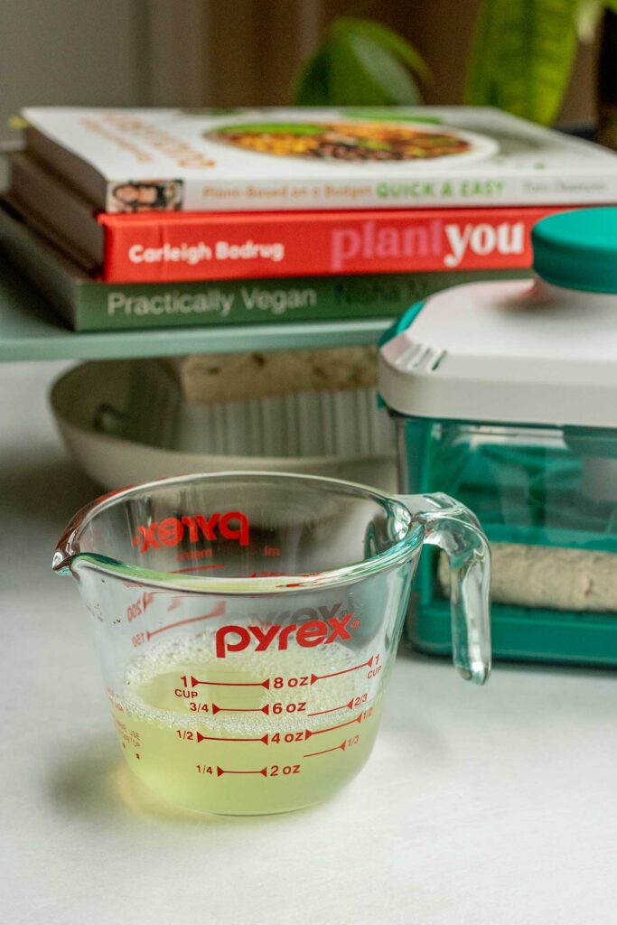 Tofu being pressed in a tofu press and a DIY tofu press with a measuring cup in front holding the pressed liquid.