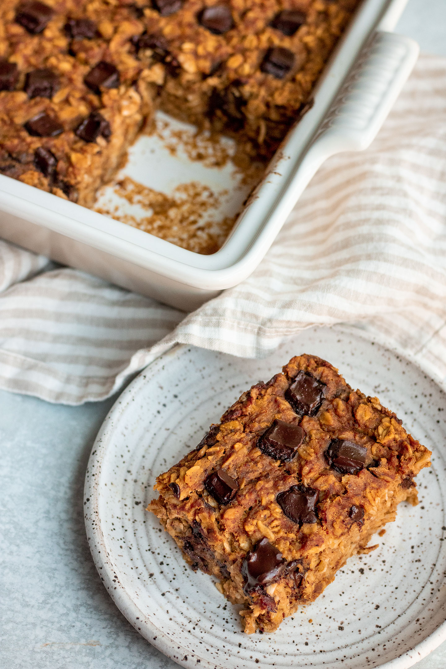 A slice of baked pumpkin oatmeal on a plate with the baking dish with remaining baked oats in the background.
