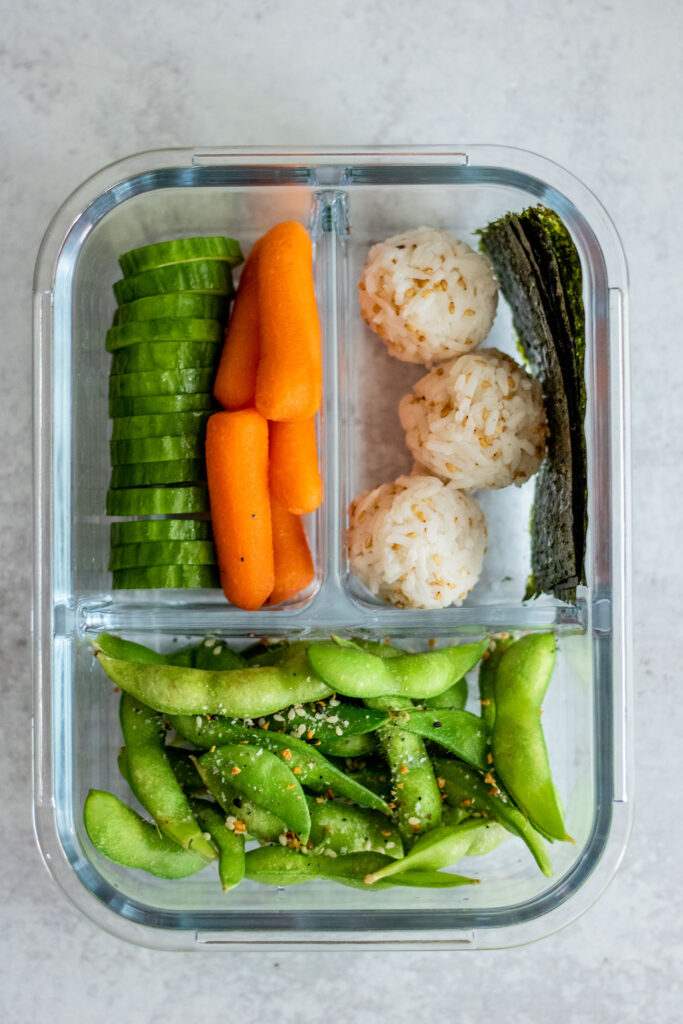 Rice ball bento box filled with cucumbers and carrots in one container, rice balls and nori in another container and edamame in a separate compartment.
