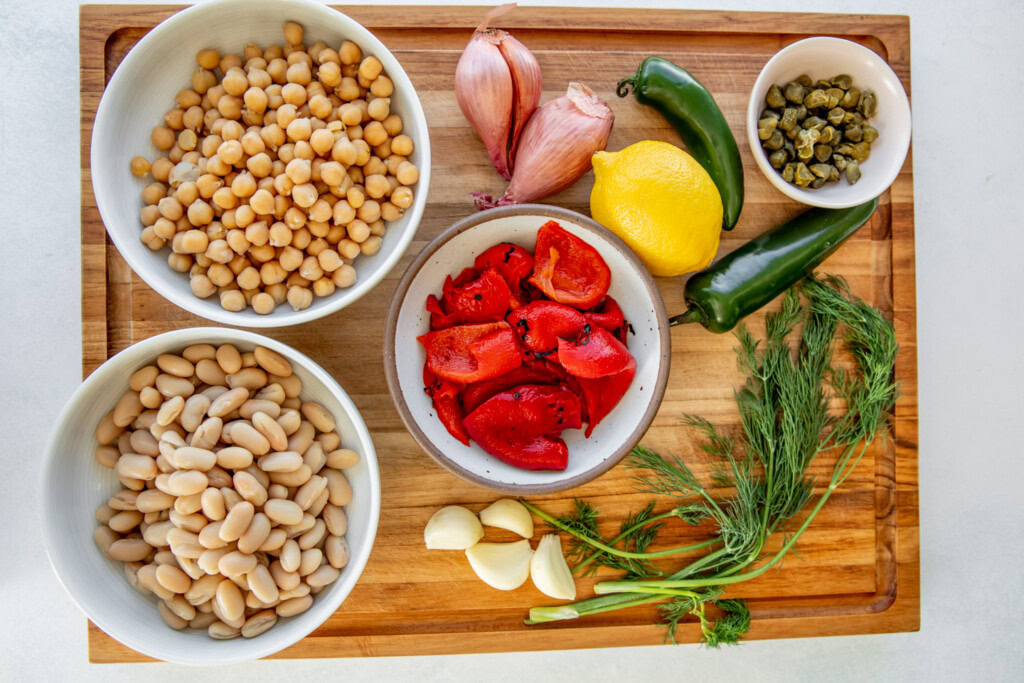 Cutting board topped with bowls of chickpeas, cannellini beans, roasted bell peppers, garlic cloves, dill, 2 jalapenos, 2 shallots, a lemon, and a bowl of capers.