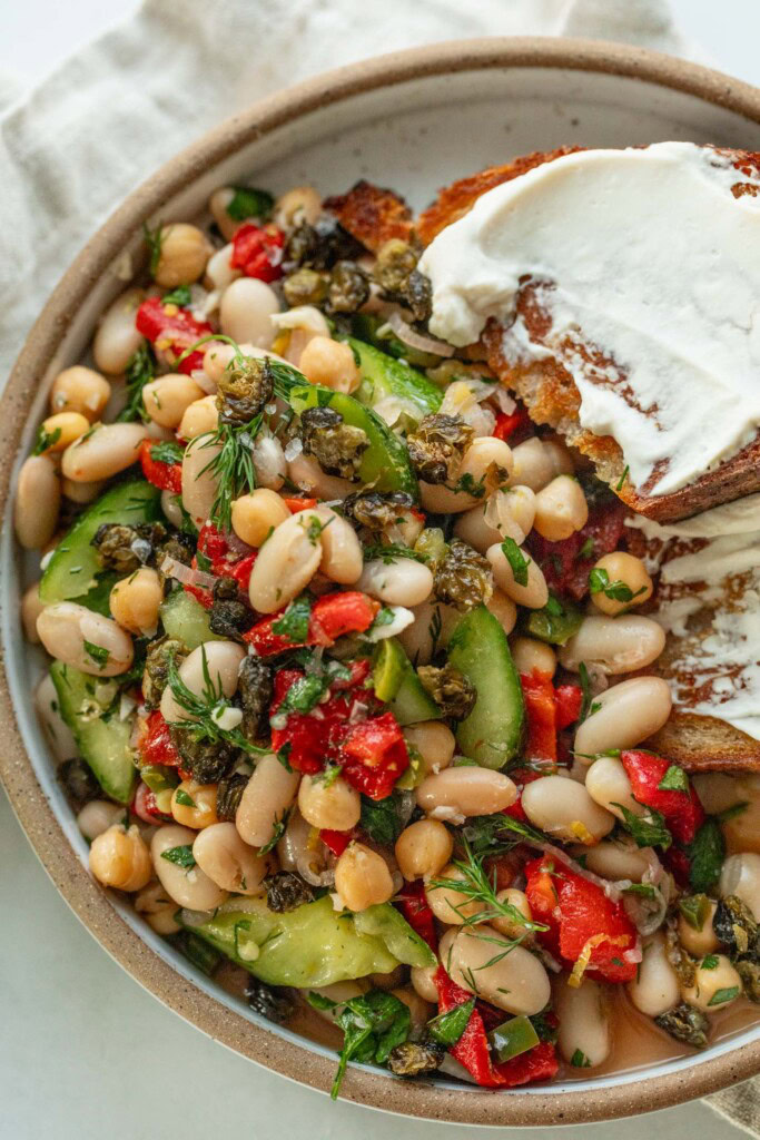 Top down view of a bowl of bean salad topped with fried capers and served with sliced toasted bread smeared with cream cheese.