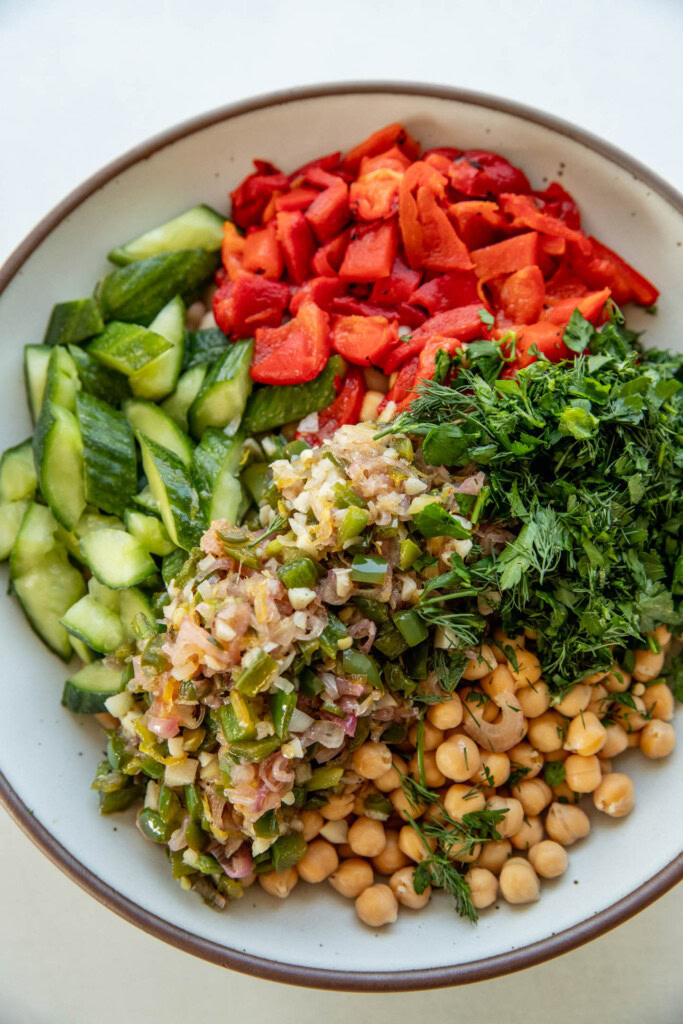 Large mixing bowl with chickpeas, white beans, dill, parsley, roasted bell peppers, smashed cucumbers, and a shallot lemon dressing.
