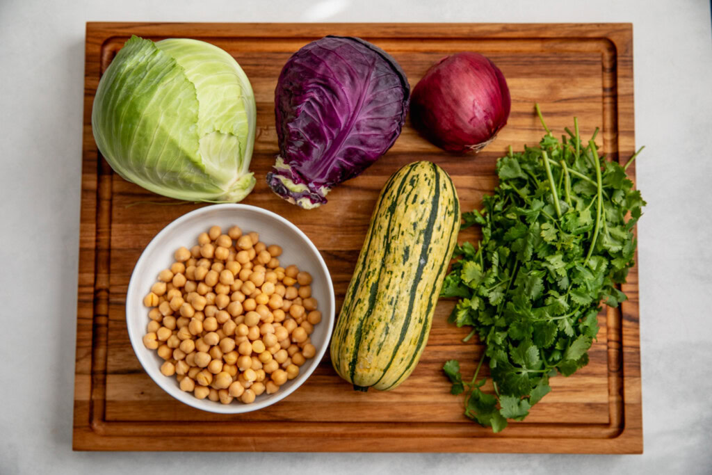 Cutting board with heads of red and green cabbage, bowl of chickpeas, delicata squash, red onion, and cilantro on it.