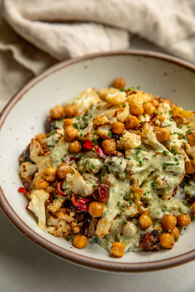 Side view of a bowl of rice topped with roasted cauliflower, dates and chickpeas then covered in an herby tahini sauce.