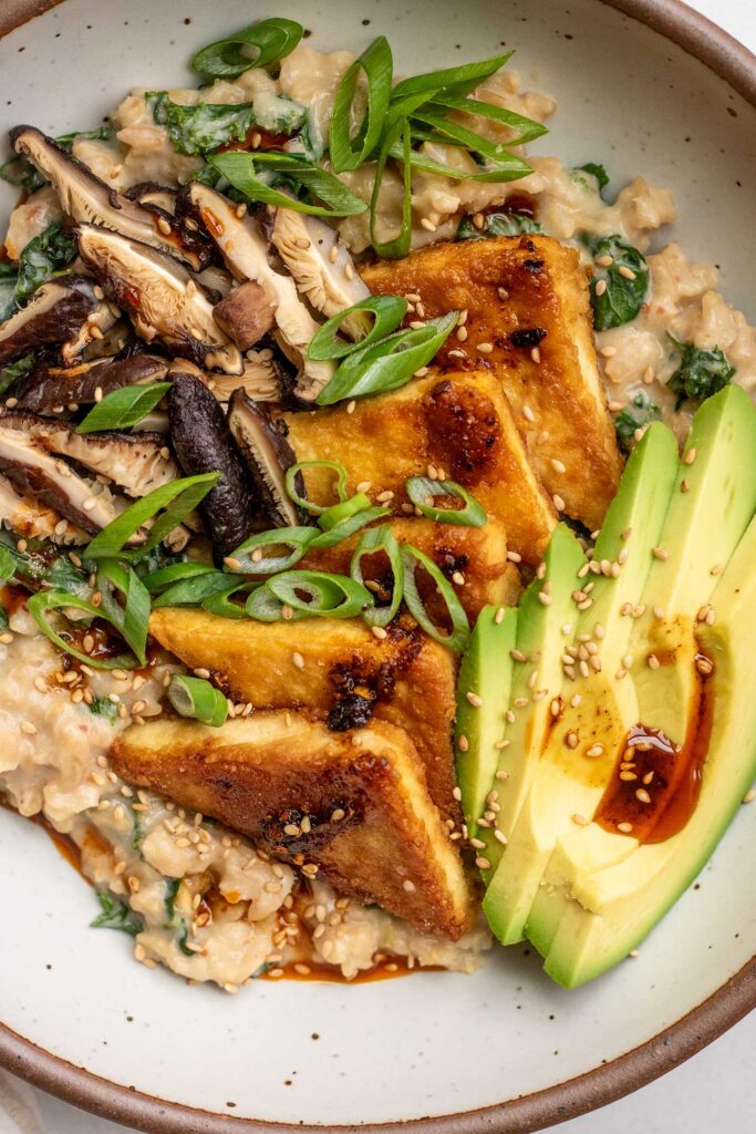 Close up of a bowl of savory oats topped with sliced avocado, pan-seared tofu, mushroom slices and scallions.
