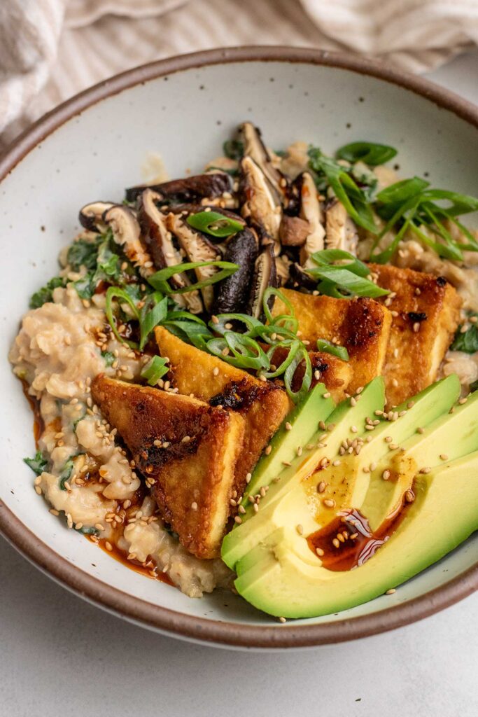 Bowl of savory oatmeal topped with pan-seared tofu, avocado, mushroom slices and scallions then drizzled with chili oil.