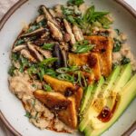 Top down view of a white bowl filled with miso oatmeal then topped with tofu triangles, avocado slices, sliced mushrooms and scallions.
