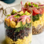 Two jars standing next to each other and loaded with tofu scramble, black beans, kale, potatoes, avocado, and pickled onions.