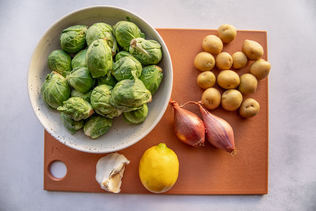 Cutting board with baby potatoes, shallots, brussels sprouts, lemon, and garlic.