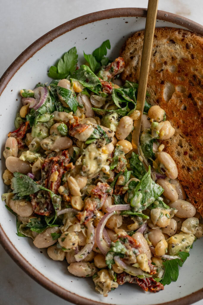 Top down of a bowl of bean salad and toasted bread being eaten with a spoon.
