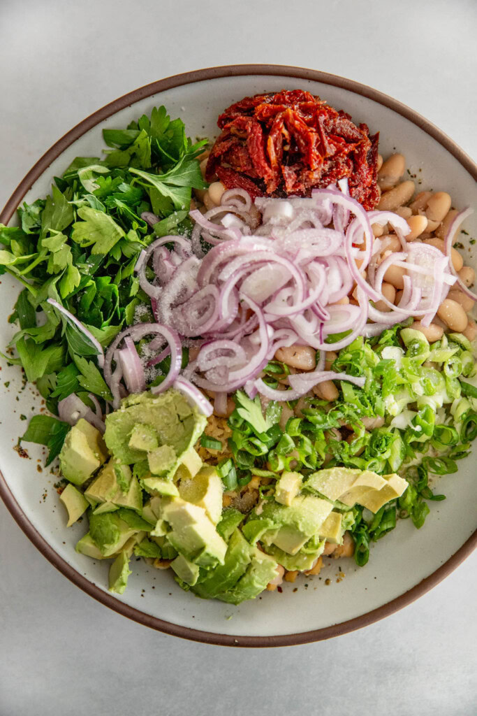 Mixing bowl filled with dressing, chickpeas, white beans, shallots, sun-dried tomatoes, avocados, cappers, scallions and parsley.