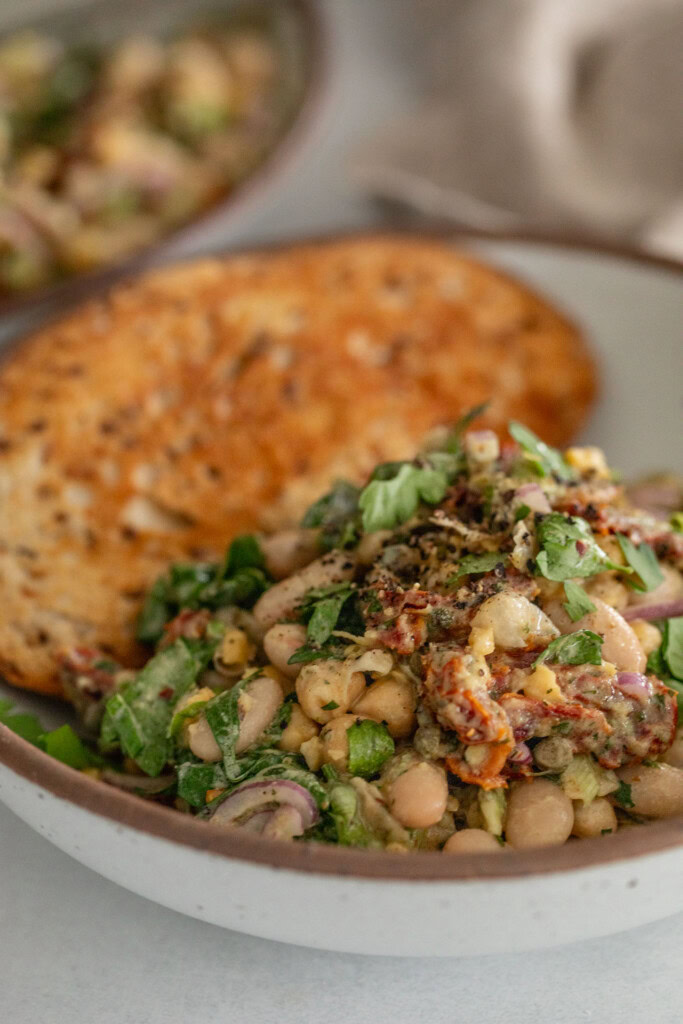 Side view of a bowl of bean salad served with a piece of toasted sourdough bread tucked into the back of the bowl.