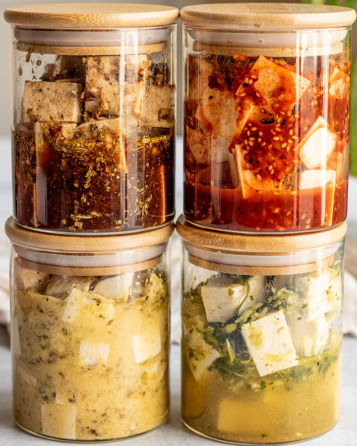 Four jars stacked on top of each other filled with 4 different marinades including balsamic tofu, gochujang tofu, tofu feta and cilantro lime tofu.