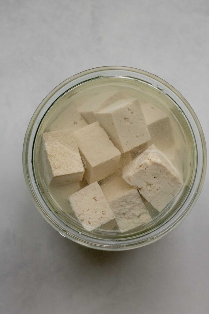 Close up view of tofu cubes soaking in a large jar with salt water.