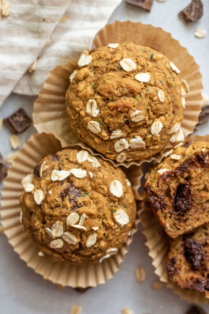 Chocolate chip zucchini muffins in wrappers with a close up of the muffins together, surrounded by chocolate chips and oats.