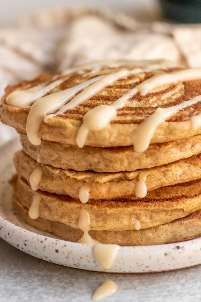 View of a stack of cinnamon roll pancakes on a white plate drizzled with frosting.