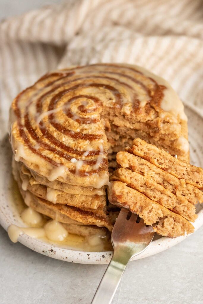 Taking a fork full of cinnamon roll pancakes from a stack on a plate.